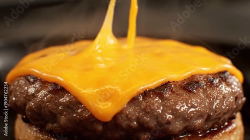 Close-up view of a sizzling burger patty being drenched with a cascade of melted cheddar cheese on a light bun, captured from a top-down kitchen perspective during cooking preparation
