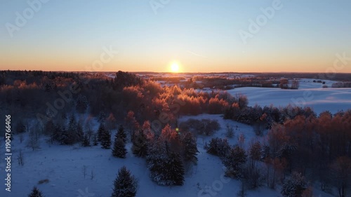 4K aerial drone flight into the sun over Lithuania winter nature at golden hour, glowing sunset above snow-covered forest, frosty trees and open fields, cinematic landscape