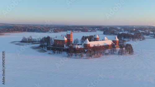4K aerial drone orbit rising clockwise around Trakai Island Castle on frozen Lake Galvė, Lithuania, highlighting red brick towers in snowy winter sunset, cinematic establishing shot