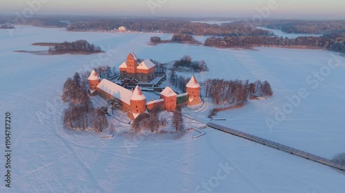 4K aerial drone ascent revealing Trakai Island Castle on Lake Galvė, Lithuania, surrounded by snow and frozen ice at pastel winter sunset, cinematic establishing shot