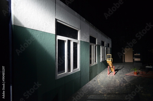 Dimly lit theatrical set with a long exterior wall, windows, and a single chair under spotlight, creating a moody, cinematic atmosphere inside a stage or rehearsal space.