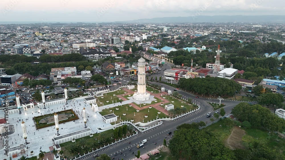 Fototapeta premium Aerial view of a grand mosque and monument in a city surrounded by lush greenery and modern buildings with tall minarets and a prominent white tower.