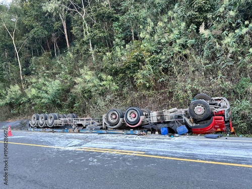 A high-impact shot showing a large red semi-trailer truck completely overturned on the side of a winding mountain road.