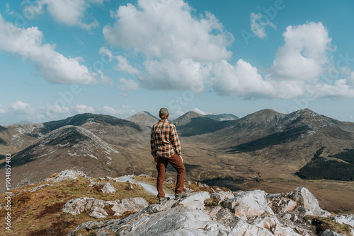 A hiker stands atop a rugged quartzite peak in Connemara National Park, surveying the dramatic Twelve Bens mountain range under a vast, cloud-filled sky.