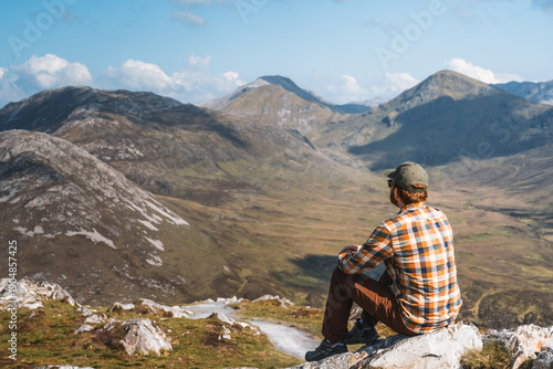 Foto A hiker sits atop a rugged quartzite peak in Connemara National Park, surveying the dramatic Twelve Bens mountain range under a vast, cloud-filled sky