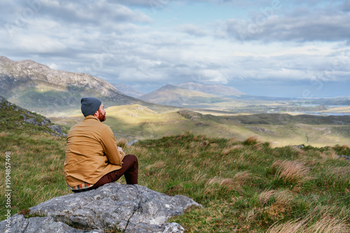 A hiker sits atop a rugged peak in Connemara National Park, surveying the beautiful Irish landscape under a vast, cloud-filled sky.