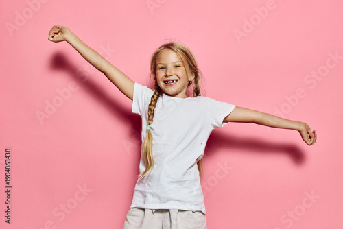 Φωτογραφία Happy child with blonde hair in braids wearing white t-shirt raising arms in celebration against pink background