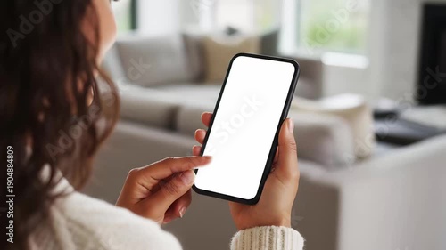 Hand holding mobile smartphone with blank white screen mockup while touching display in modern living room interior background for app design website presentation display copy space
