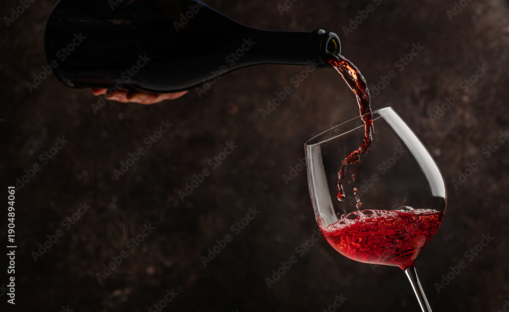 Fototapeta premium Pouring red wine into the glass on rustic dark background. Pour alcohol, winery concept.