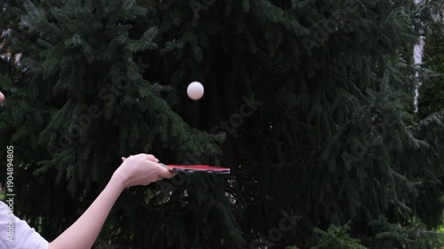 Close-up of a hand practicing table tennis with a red paddle, hitting a white ball against dense green trees. Captures precision, focus, and repetitive training motion in an outdoor setting.