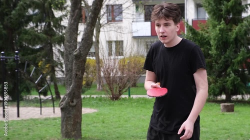 Dynamic outdoor table tennis match in a sunny park. Players actively hitting the ball with rackets on a concrete table, green grass and playground visible in the background. Captures fun, competitive 