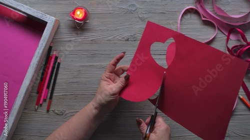Top view of hand cutting heart shapes from red cardboard with scissors. DIY handmade Valentine Day card preparation and creative craft process