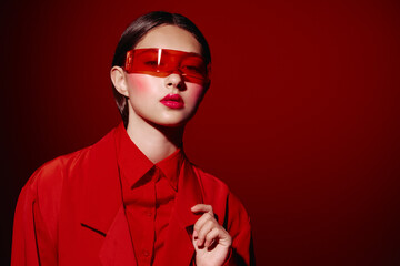 Stylish woman in bold red fashion outfit poses against a red background wearing futuristic tinted sunglasses. Beauty makeup highlights glossy lips and well-defined cheeks. Creative concept featuring