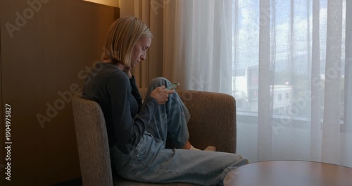 Woman sits in a chair in hotel room, she is focused on her phone and looks relaxed.