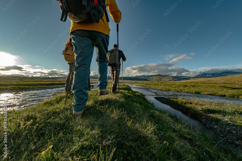 Obraz premium Two hikers with backpacks and trekking poles are walking along a narrow grassy ridge beside a river in a vast, green mountain landscape under a bright, sunny sky.