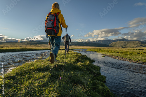 Valokuva A man and a woman in outdoor gear with backpacks and trekking poles are actively hiking along a grassy river bank on a sunny day in a vast mountain landscape