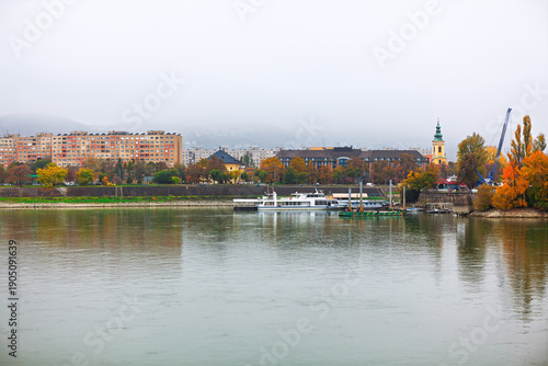 Danube River in Budapest, Hungary, looking towards the Buda side hilly area from the flatter Pest riverfront. Buildings on the bank include a prominent church steeple with boats moored along shore