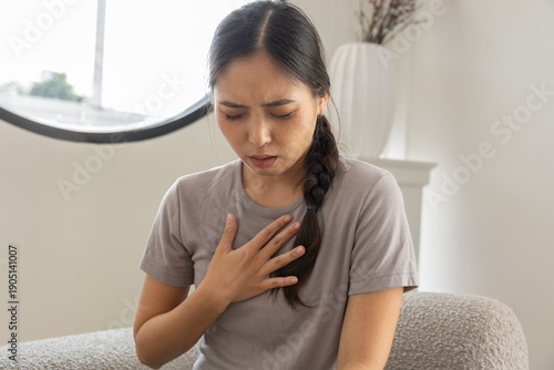Young distressed Asian woman feeling sharp chest pain breathing hard suffering heart attack or acid reflux symptoms while sitting home alone unwell sick female patient clutching chest having