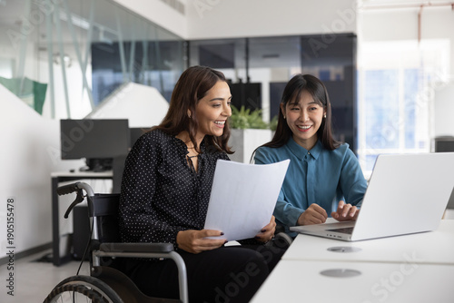 Happy female Middle Eastern office employee with disability consulting young Chinese colleague. Diverse coworkers of inclusive company working together at laptop, typing, smiling