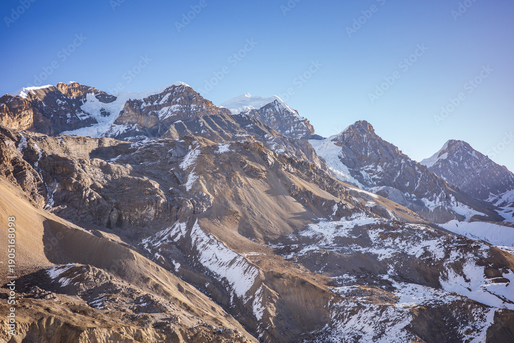 Obraz premium Dramatic view of rugged Himalayan mountain peaks with scattered snow and exposed rocky slopes under a clear blue sky. The raw alpine terrain and sharp ridgelines convey power, scale, and endurance