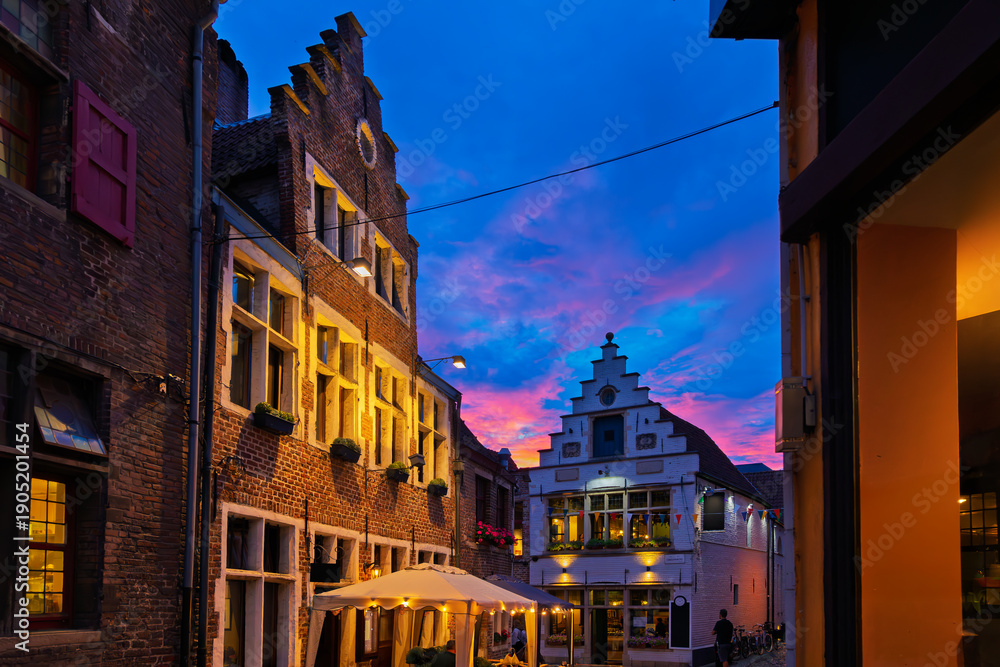 Fototapeta premium Illuminated traditional Belgian gabled houses and cafes on a charming, narrow, cobbled alley in the historic Patershol district of Ghent Belgium at twilight.