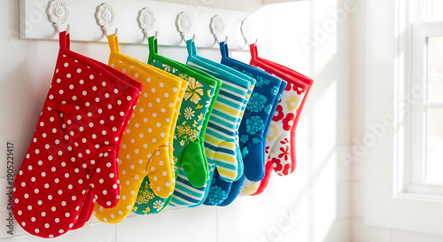 Colorful oven mitts hanging on a white rack against a white wall near a bright window