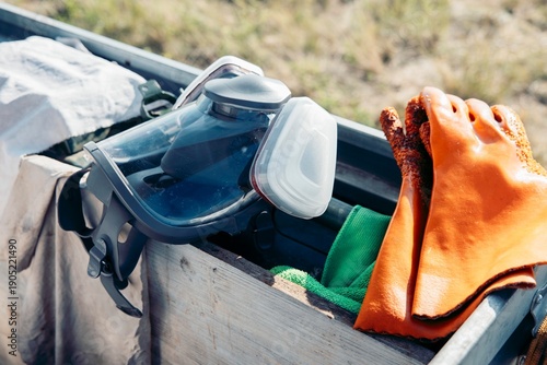 protective equipment stored in wooden box outdoors, safety goggles respirator and orange work gloves arranged, field work in natural environment, concept of construction, industry, safety
