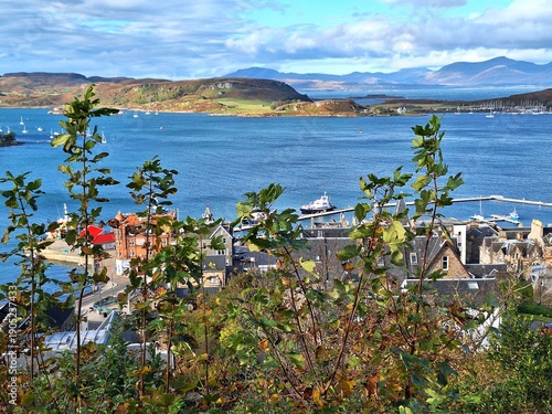 Картината върху платно Panoramic view over Oban Bay in Argyll and Bute, Scotland, captures blue waters dotted with boats and yachts, rolling hills in the distance, and traditional stone buildings clustered along shoreline