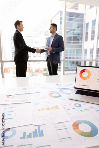 Conclusion of successful negotiation, mutual agreement, trust. Close up of laptop view with displaying pie charts data and papers spread out on table, two businessmen shaking hands stand in background
