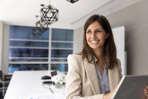 Smiling middle-aged 40s Caucasian businesswoman looking into distance, distracted from tablet usage, planning strategy, thinks on new career opportunities. Business vision, aspirations and success