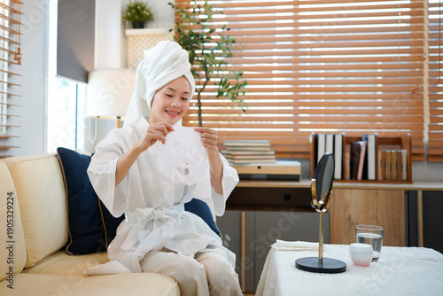 Asian female in white bathrobe putting sheet mask on face with help of makeup mirror with natural skincare remedies. Pretty woman pampering herself with beauty treatment