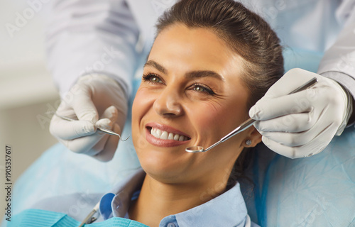Happy woman smiling during routine checkup in dental clinic, dentists with tools checking teeth. Confident healthy smile of patient receiving examination, doctors hands holding mirror and scaler