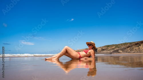 Beautiful middle-aged woman model sitting on sandy beach in summertime. Side view. Monte Clerigo beach on Algarve coast in Portugal
