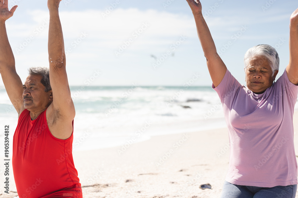 Fototapeta premium Senior couple exercising together with arms raised at beach against sky on sunny day
