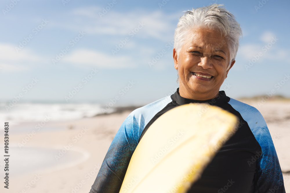 Fototapeta premium Portrait of smiling senior woman carrying surfboard at beach against sky on sunny day