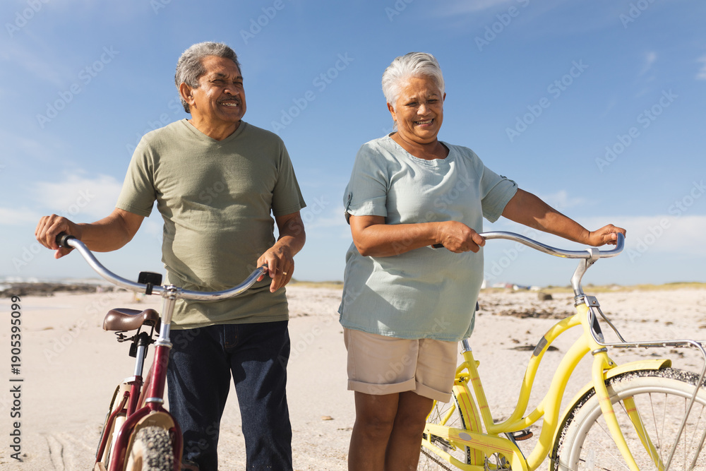 Fototapeta premium Happy multiracial senior couple standing with bicycles at beach against sky during sunny day