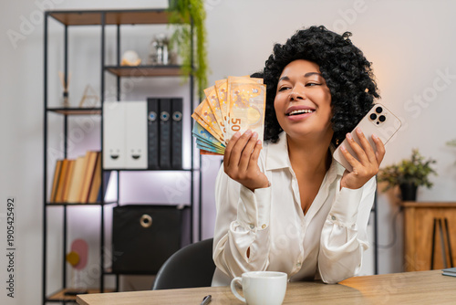 African American businesswoman at home office laughs holding smartphone surprised by profit alert counts euro cash. Freelancer girl at table plans spending smiling imagining travel fun retail dreams