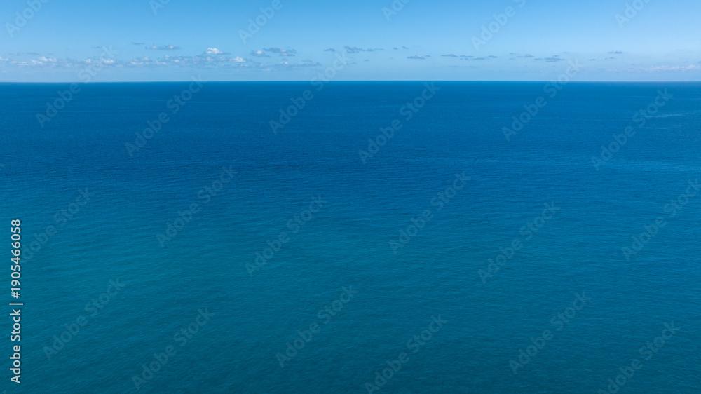 Obraz premium A serene wide shot of the deep blue Sea, with a clear horizon line under a soft blue sky and distant faint clouds. Aerial view of the vast ocean.