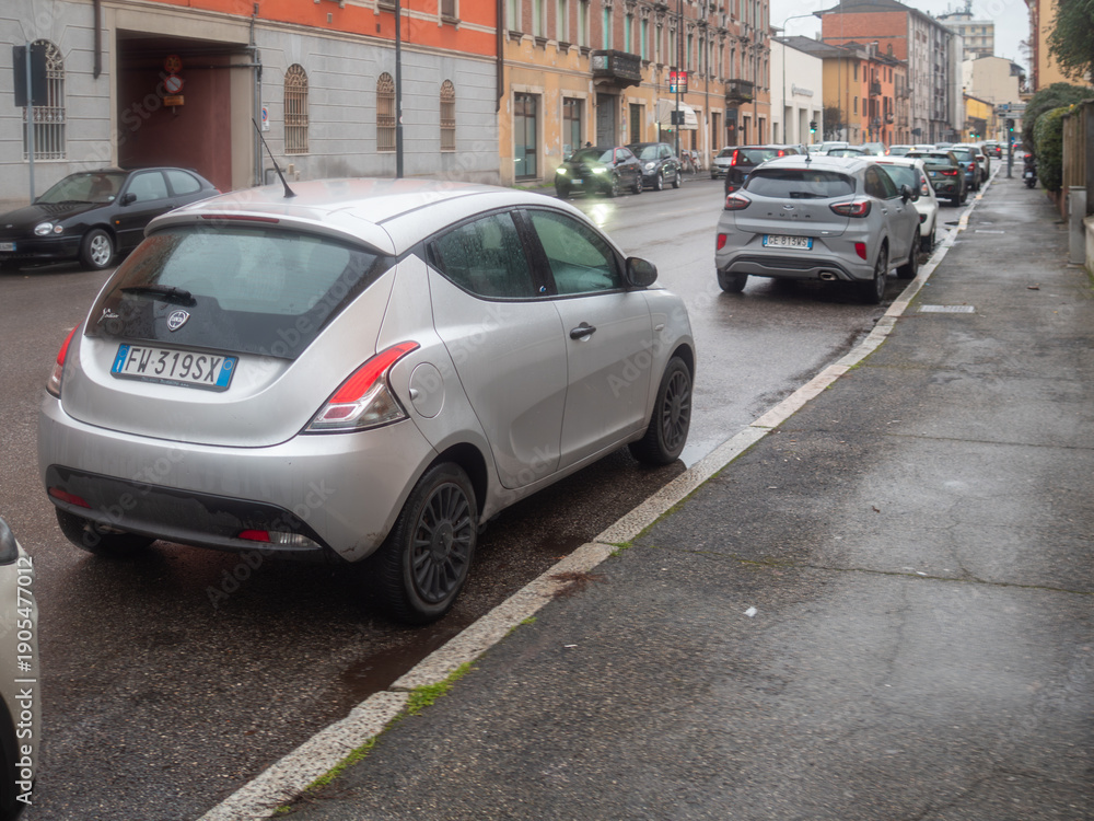 Naklejka premium Silver Lancia Ypsilon and Ford Puma navigating wet city street during a rainy day traffic commute in Italy
