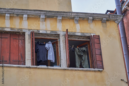 Clothes hanging to dry on ropes across a window with open brown wooden shutters on an old yellow wall in Venice.