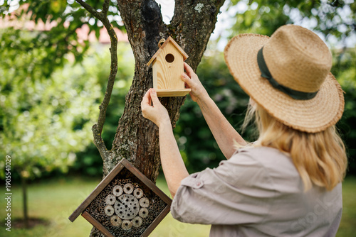 Woman gardener is placing wooden birdhouse and insect house on apple tree for supporting biodiversity in garden