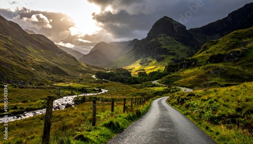 Road winds through a sunlit valley with rolling green hills under a dramatic sky, and a river