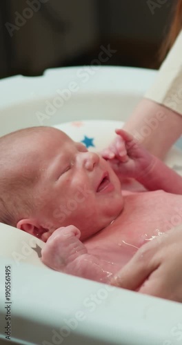 A distressed newborn baby cries loudly while being bathed in a small tub, with hands gently holding its body