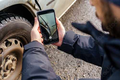 Person taking a photo of car damage with a smartphone. Scratches and dents are visible on a silver car fender, showing vehicle condition after a minor accident.