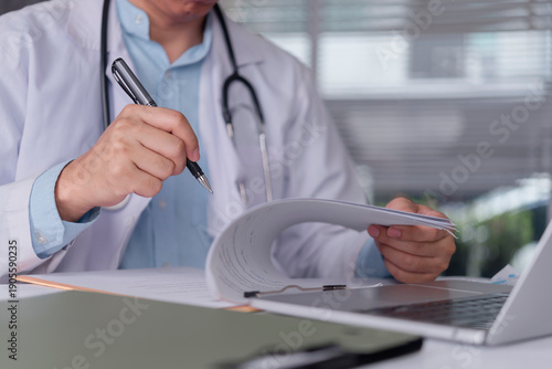 Foto doctor hand writing medical report on clipboard and reviewing patient document at hospital office with laptop