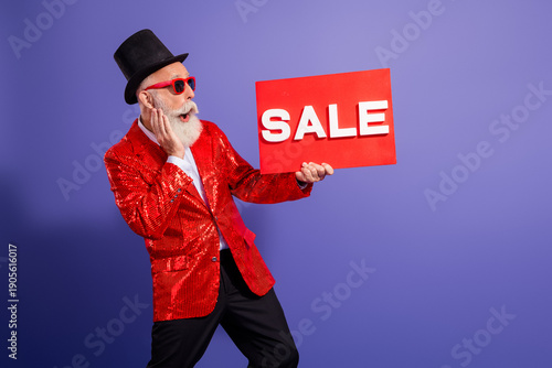 Bearded performer in a red sequin jacket holds a bold sale sign during a festive showman moment against a vivid purple backdrop
