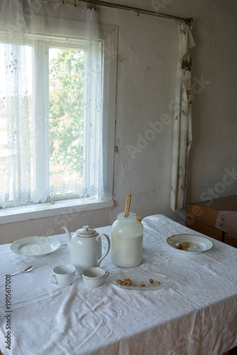 Traditional breakfast table setting with white dishes, teapot, and pitcher by a bright window in a rustic room