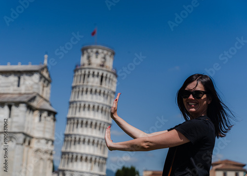 Woman posing in front of leaning tower creating optical illusion on city square.