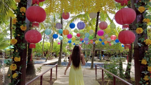 Asian girl in yellow dress walks in nature and passes under a bridge full of typical colorful Thai lanterns -  coconut palms, oriental lanterns and a travel atmosphere