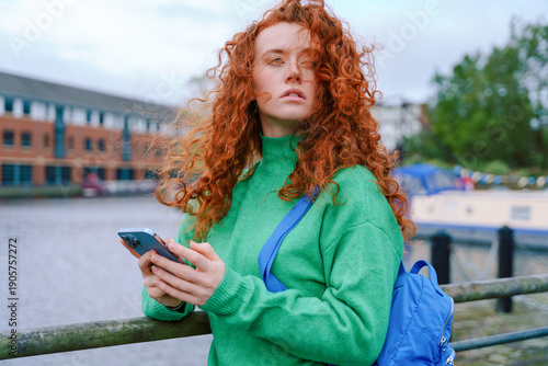Photos Young woman with curly red hair uses smartphone by the canal in a city on a clou